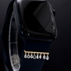 Puede incluir: Un Apple Watch negro con una banda dorada y un dije colgante con cristales transparentes en forma de lágrima.