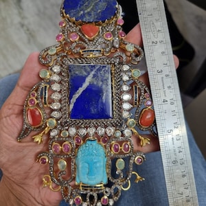 May include: A gold pendant with a blue lapis lazuli stone, a turquoise Buddha, and red and pink gemstones. The pendant is surrounded by a silver filigree design and is approximately 2 inches in length.