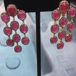 Op de afbeelding: Een paar oorhangers met roze edelstenen en een halo-setting van diamanten. De oorhangers staan op een zwarte displaystandaard.