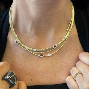 Puede incluir: Un collar de cadena de oro con tres hebras y pequeñas piedras preciosas rojas. El collar lo lleva una persona con una camisa negra.