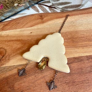 May include: A cream-colored, cloud-shaped ceramic ornament featuring a central amber-toned teardrop crystal. Two small, dark metal bee charms hang from the bottom edges on thin chains, with a matching chain attached to the top for hanging.