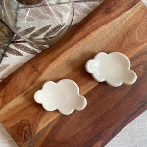 May include: Two small, off-white ceramic dishes shaped like stylized clouds. Each dish features a smooth, glossy finish with rounded, scalloped edges, designed as a shallow tray for holding small items. The pieces are solid in color and uniform.