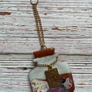 May include: A ceramic pendant in the shape of a potion bottle, featuring a gold chain and a rectangular charm. The bottle has a white and reddish-brown glaze. The charm has an engraved design. The pendant hangs from a gold ring.