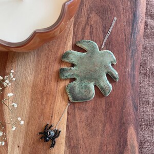 May include: A hanging ornament featuring a glossy, mottled green ceramic leaf shape suspended by a silver-toned chain. Attached to the bottom of the leaf is a small, beaded spider with black legs and a round, black and white striped body.