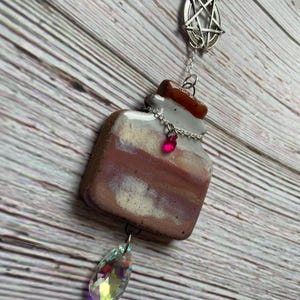 May include: A handcrafted, square-shaped pendant with a potion bottle design. The pendant features a brown, white, and pink color scheme, a silver chain, a pink bead, and a large, iridescent crystal. A silver pentagram charm hangs above.