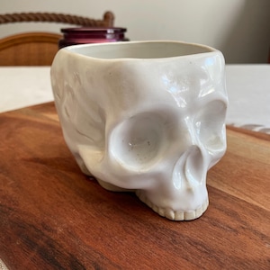 May include: A white ceramic skull-shaped planter or container with a glossy finish. The object features hollow eye sockets, a defined nasal cavity, and visible teeth along the lower jaw. The top is open, revealing a smooth interior surface.