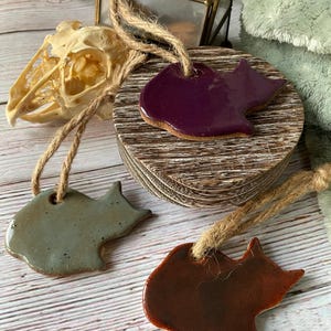 May include: Three ceramic cat-shaped ornaments with glossy glazes in purple, sage green, and reddish-brown. Each ornament features a hole at the top with a natural jute twine loop for hanging, showcasing smooth, reflective surfaces and distinct silhouettes.