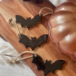May include: Three bat-shaped ornaments made of dark, glossy ceramic, each featuring a jute twine loop for hanging. Two bats have a solid black finish, while the middle bat displays a black surface with white speckled details throughout.