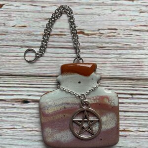 May include: A ceramic pendant in the shape of a bottle with a silver chain and a pentagram charm. The pendant has a white, brown, and pink color scheme. The chain is attached to a silver ring.