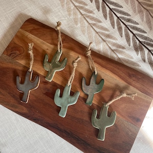 May include: Five ceramic cactus ornaments in varying shades of green and brown, each featuring a smooth, glazed finish and a rustic jute twine loop attached at the top for hanging. The ornaments are shaped like saguaro cacti with two arms.