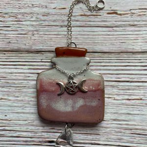 May include: A ceramic pendant in the shape of a potion bottle, with a silver chain and a clear crystal drop. The bottle has a gray top, a reddish-pink middle, and a silver crescent moon and pentacle charm.