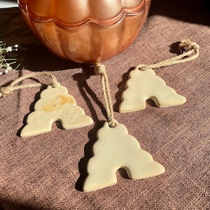 May include: Three off-white, arch-shaped ceramic ornaments with smooth surfaces, each featuring a hole at the top threaded with a natural jute twine loop for hanging. The ornaments have a tiered, stepped design and are arranged in a cluster.
