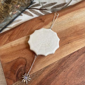 May include: A white ceramic spider web ornament with scalloped edges, suspended by a silver-toned metal chain. A small silver-toned spider charm with a central iridescent purple stone hangs from the bottom of the web, creating a decorative piece.