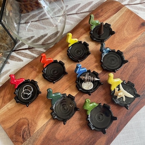 May include: A collection of nine small, circular black ceramic dishes, each featuring a unique, colorful dinosaur-shaped figurine attached to the rim. The dinosaurs are painted in shades of red, yellow, green, and blue. Some dishes hold small items.