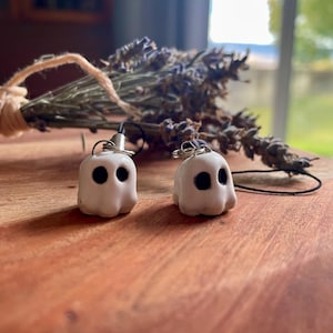 May include: Two small, white, ghost-shaped charms with smooth surfaces and simple black circular eyes. Each charm features a silver-toned metal loop at the top, attached to a thin black cord, designed for hanging or as a decorative accessory.