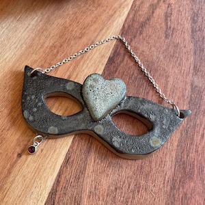 May include: A ceramic masquerade mask ornament featuring a dark, speckled glaze with a textured, light green heart centered between the eye openings. A silver-toned metal chain is attached to the top, and a small, round, deep red gemstone charm hangs from the lower left side.