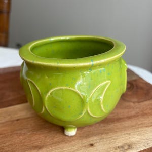 May include: A lime green ceramic cauldron with a glossy finish and blue speckles. The exterior features a raised triple moon symbol embossed on the front. This small, rounded vessel stands on three short feet and has a wide, open circular rim.