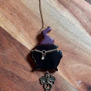 May include: A black cauldron ornament with a purple smoke-like top, suspended by a gold-toned chain. A gold chain wraps around the cauldron with a small clear crystal charm, and a bronze-colored dragon pendant hangs from the bottom of the piece.