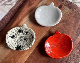 May include: Three small ceramic dishes shaped like pumpkins. One dish is plain white, one is bright orange with black speckles, and one is cream-colored with a black spiderweb pattern and several black spiders printed on the surface.