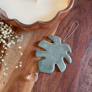 May include: A sage green ceramic monstera leaf ornament with a speckled finish, featuring a silver-toned metal chain for hanging and a clear, faceted crystal drop attached to the bottom point of the leaf, creating a decorative hanging accent.