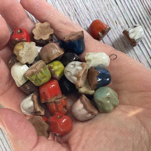 May include: A handful of small, ceramic acorn-shaped beads in various colors, including red, white, blue, and green. The beads have a glossy finish and are arranged in a person's open palm. The background is a light-colored wooden surface.