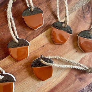 May include: A set of six ceramic acorn ornaments, each featuring a glossy orange body and a dark brown textured cap. Each ornament is attached to a thick, natural-colored jute twine loop for hanging, creating a uniform collection of autumn-themed decor.