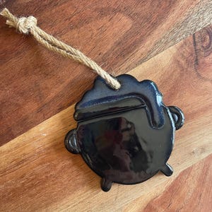 May include: A glossy black ceramic cauldron ornament featuring a rounded body with three small feet and two side handles. A thick, textured drip of dark blue glaze runs down from the rim. The piece is suspended by a natural tan jute twine loop.
