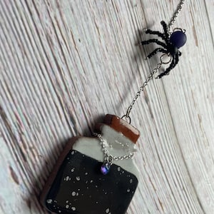 May include: A handcrafted Halloween-themed charm featuring a small, dark potion bottle with a brown cork and a silver chain. A black beaded spider with a purple bead hangs above the bottle, connected by a chain.