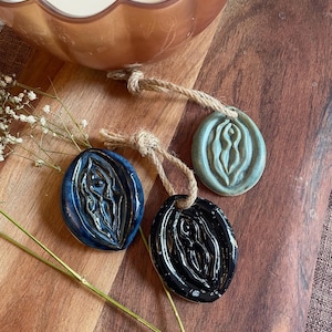 May include: Three oval ceramic ornaments with a glossy finish in blue, black, and sage green. Each piece features a raised, symmetrical organic relief design and is attached to a short, natural-colored jute twine loop for hanging purposes.