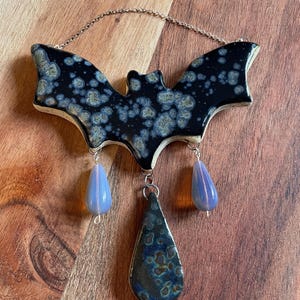May include: A ceramic bat ornament with a black and blue crystalline glaze finish. The bat features a thin silver-toned metal chain for hanging and three dangling charms: two small translucent light blue teardrop beads and one larger ceramic teardrop pendant with a matching crystalline glaze.