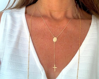 Puede incluir: Un collar dorado de capas con un pequeño colgante que presenta un icono religioso y una cruz. El collar lo lleva una persona con el pelo largo y castaño.
