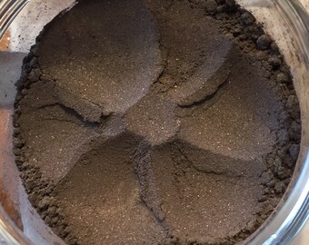 Op de afbeelding: Een heldere glazen pot gevuld met een donkergrijs, fijn gemalen poeder. Het poeder heeft een licht glinsterend uiterlijk en is in een cirkelvormig patroon gerangschikt. De pot wordt van bovenaf bekeken, waarbij de textuur en kleur van het product zichtbaar zijn.