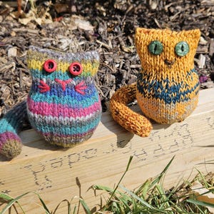 Könnte beinhalten: Zwei handgefertigte gestrickte Katzenfiguren. Eine Katze ist gestreift mit roten Knopfaugen und einer rosa gestickten Nase. Die andere ist orange mit grünen Knopfaugen und einem blau-marinefarbenen Muster.