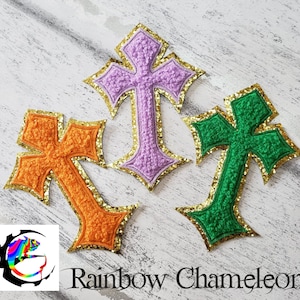May include: Three embroidered crosses with gold glitter trim. The crosses are in orange, green, and purple.