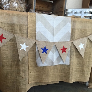 May include: A burlap table runner with a white chevron pattern and a burlap banner with red, white, and blue stars. The banner is strung across the table runner.
