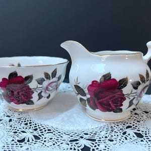 May include: A white porcelain creamer and sugar bowl set featuring a floral pattern of deep red and pale pink roses with green leaves. Both pieces have scalloped edges and are finished with a thin gold trim along the rims and handles.
