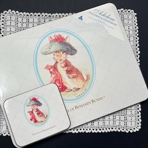 May include: A placemat and coaster set with a rabbit illustration wearing a hat and holding an apple. The set includes a rectangular placemat and a square coaster. The text "The Tale of Benjamin Bunny" is visible. The set is made by Ashdene.