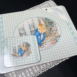 May include: A placemat and coaster set with a Peter Rabbit design. The set includes a rectangular placemat and a square coaster. Both items have a light blue and white checked pattern with a Peter Rabbit image in the centre.