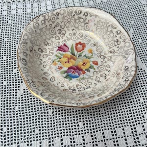 May include: A small, cream-coloured ceramic dish with a gold rim and a floral cross-stitch design. The dish has a delicate, scalloped edge and a gold floral pattern on the surface.