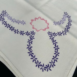 May include: A white fabric cloth featuring hand-stitched embroidery. The design includes a central pink scalloped flower shape surrounded by a U-shaped garland of dark purple petals, with additional light purple floral accents on the upper sides.