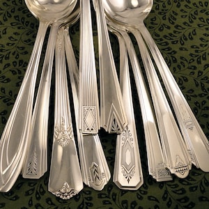 May include: A set of ten silver spoons with ornate designs on the handles. The spoons are arranged in a fan shape, with the handles pointing towards the top of the image.