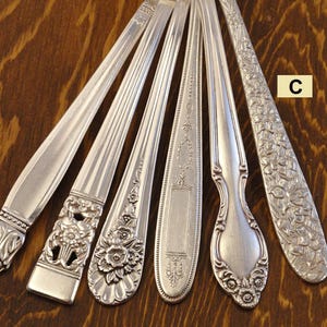May include: A collection of antique silver spoons and forks, each with unique decorative patterns. The silverware features ornate floral and geometric designs, showcasing various styles and eras. The spoons and forks are arranged on a dark wooden surface.