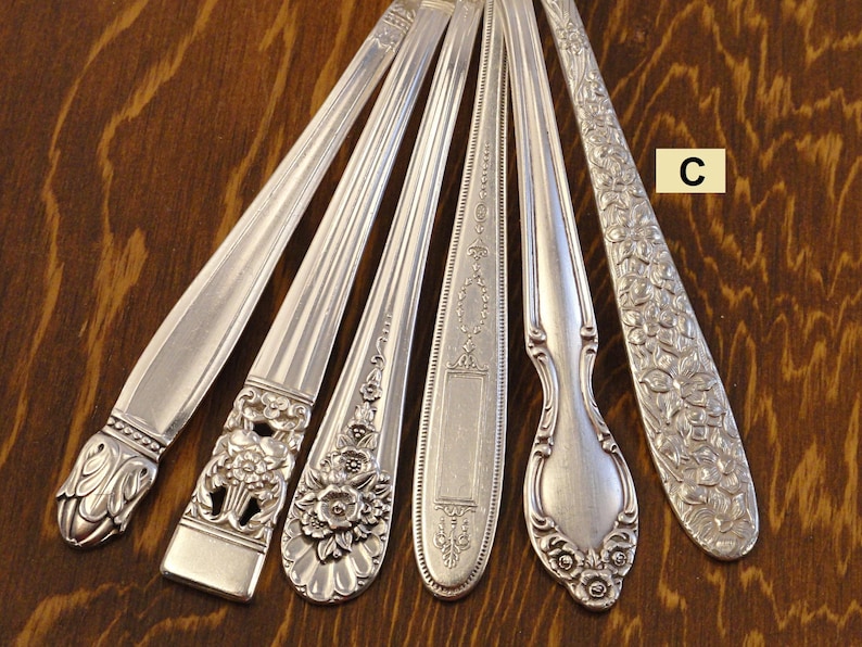 May include: A collection of antique silver spoons and forks, each with unique decorative patterns. The silverware features ornate floral and geometric designs, showcasing various styles and eras. The spoons and forks are arranged on a dark wooden surface.