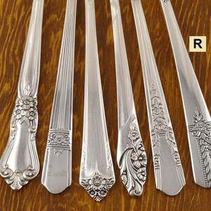 May include: Six silver spoons with different handle designs are arranged on a wooden surface. The handles feature floral and geometric patterns. The spoons are made of a shiny, reflective metal.