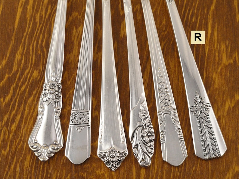 May include: Six silver spoons with different handle designs are arranged on a wooden surface. The handles feature floral and geometric patterns. The spoons are made of a shiny, reflective metal.