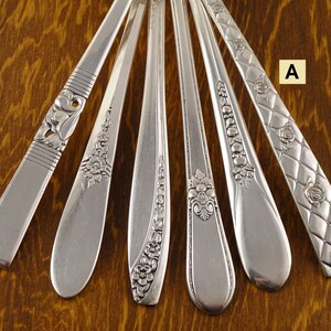 May include: An assortment of silver-colored flatware, including spoons and forks, with various decorative handle designs. The handles feature floral and geometric patterns. The flatware is arranged on a wooden surface.