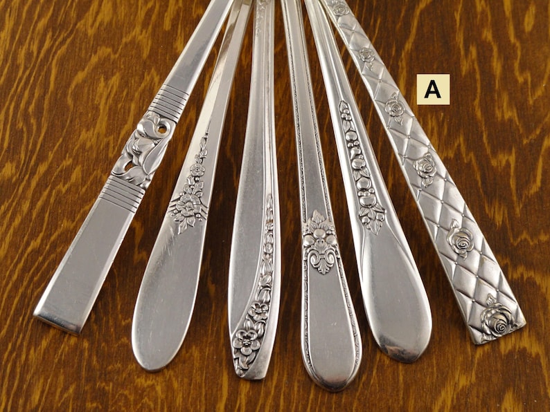 May include: An assortment of silver-colored flatware, including spoons and forks, with various decorative handle designs. The handles feature floral and geometric patterns. The flatware is arranged on a wooden surface.