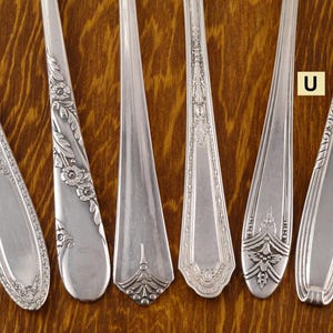 May include: Close-up of six silver spoons with ornate designs. The spoons have different patterns, including floral and geometric motifs. The spoons are arranged on a wooden surface, showcasing their intricate details and vintage style.