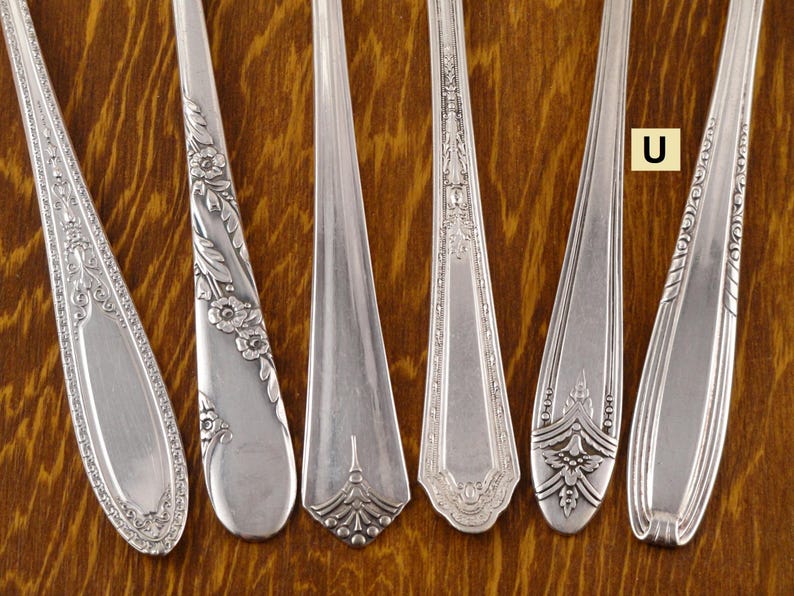 May include: Close-up of six silver spoons with ornate designs. The spoons have different patterns, including floral and geometric motifs. The spoons are arranged on a wooden surface, showcasing their intricate details and vintage style.