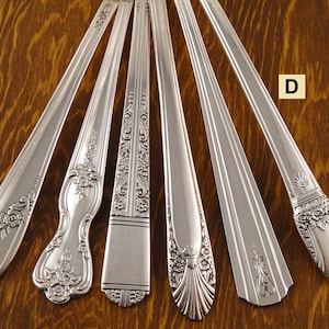 May include: Six silver-colored antique spoons arranged on a wooden surface. Each spoon features a unique decorative handle design, including floral and geometric patterns. The spoons are arranged in a fan shape, showcasing their varied styles.